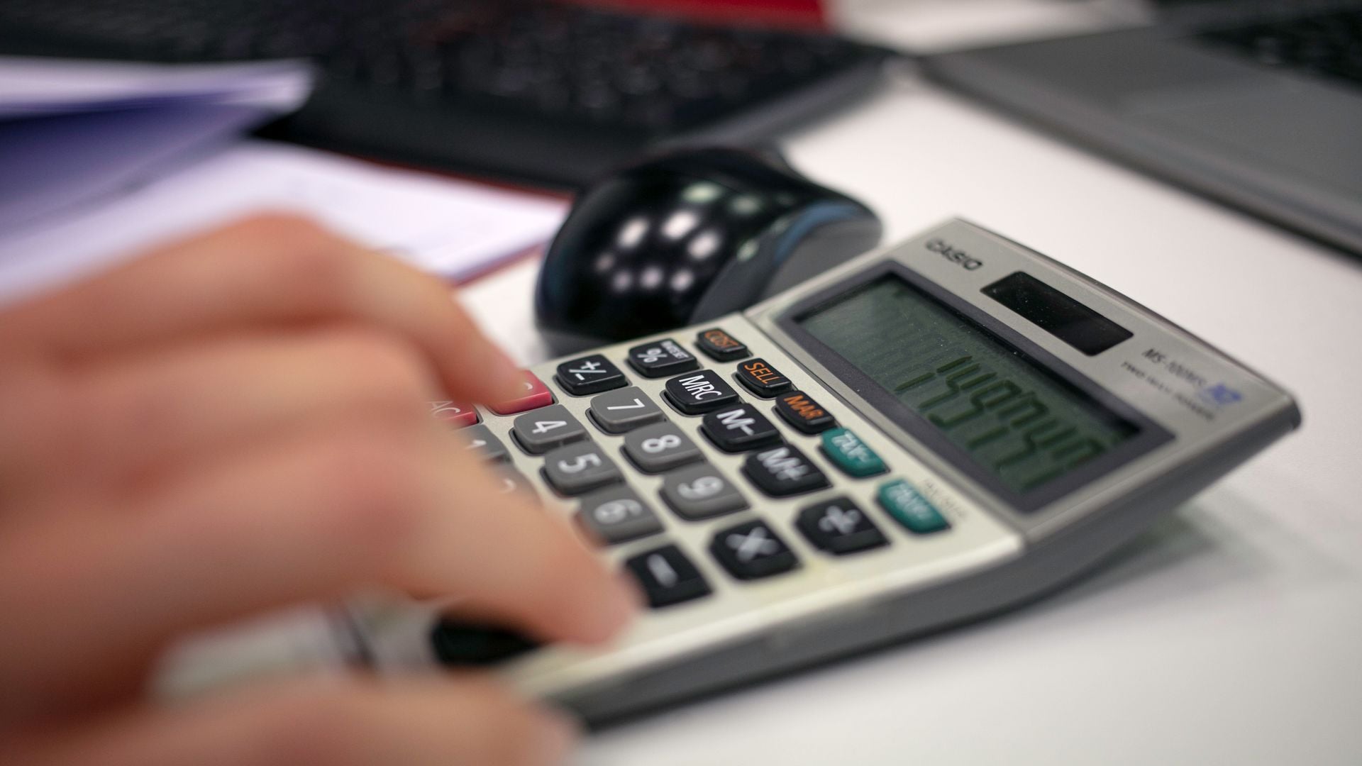 A hand using a desktop calculator beside a mouse and laptop