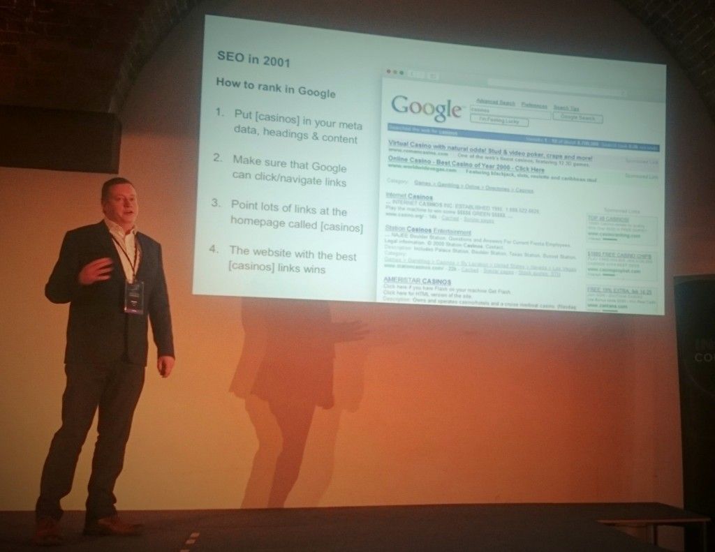 Rob Kerry stands next to a projection screen at Ayima Insights conference. The projector reads 'SEO in 2001: How to rank in Google' with a set of numbered instructions below for SEO optimising a casino website. 1. Put "casinos" in your meta data, headings and content. 2. Make sure that Google can click/navigate links. 3. Point lots of links at the homepage called "casinos". 4. The website with the best "casinos" links wins. To the right there is a screenshot of 2001-style Google search results for the word "casinos".