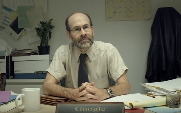 A grumpy, balding middle-aged white man with a beard and glasses sits behind a desk with his hands folded. A nameplate on the desk reads "Google".