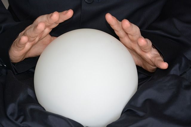 A pair of hands hovering above a large white crystal ball, which is surrounded by black cloth.