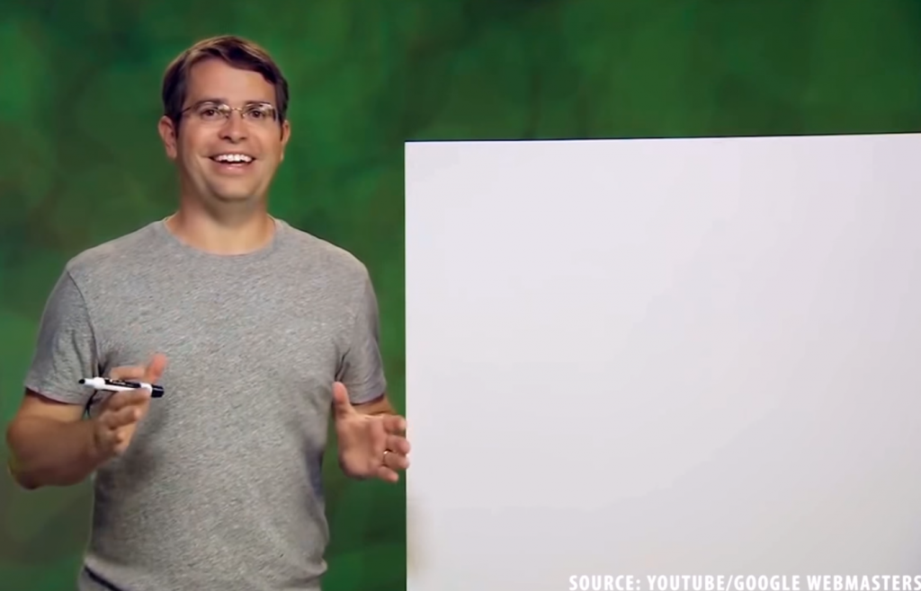 Still from SEO: The Movie showing Matt Cutts holding a whiteboard marker next to a blank whiteboard, mid-explanation of a concept. The credit in the bottom right corner reads 'Source: YouTube/Google Webmasters'.